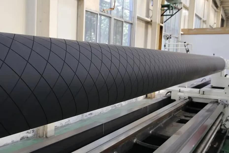 Top view of a custom industrial carbon fiber roller shaft featuring a deep diamond-patterned surface for traction, designed for high-speed film converting and printing machinery on a factory floor.